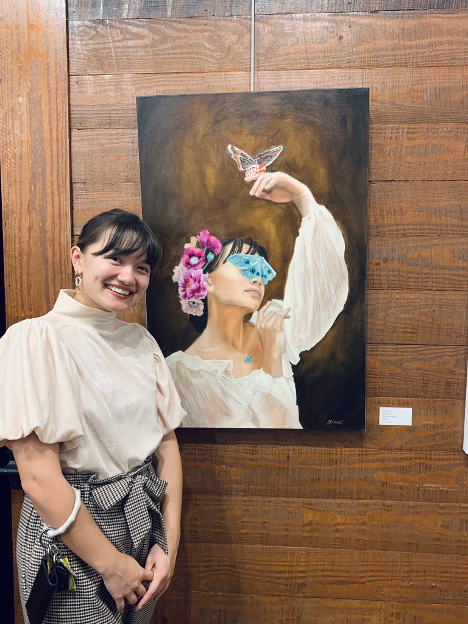Abigail standing in front of her painting 'Jade' which depicts a woman holding a butterfly and has a butterfly over her eyes and flowers in her hair.