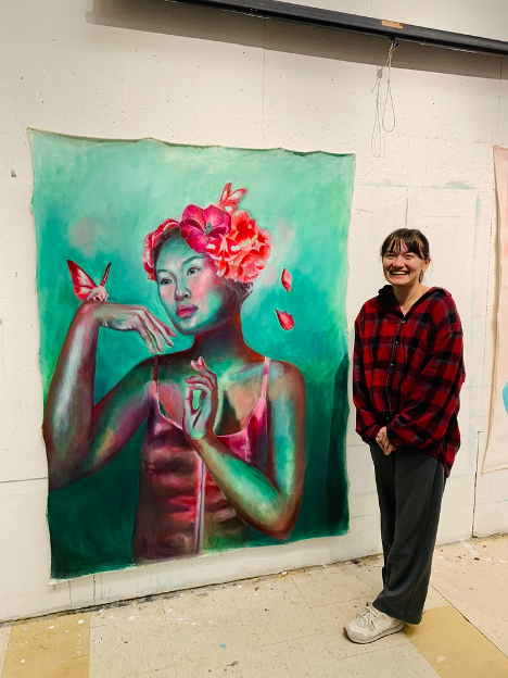 Image of Abigail standing next to her painting "Eyes to see beauty" which depicts a woman with flowers in her hair and a butterfly on her hand. The image was in the West Alabama Juried Art Show at the Dinah Washington Cultural Arts Center in Tuscaloosa
