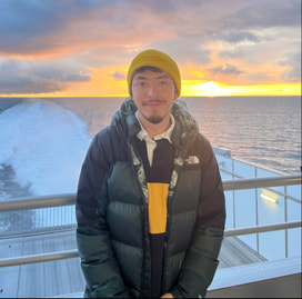 Image of Noah Jacobs, standing in a winter jacket and hat in front of the ocean while on a boat.
